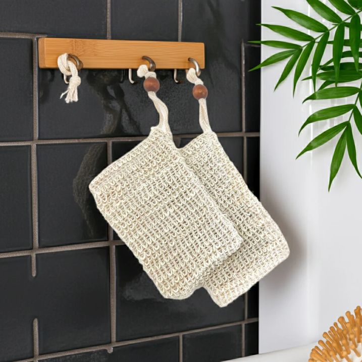 Two beige soap saver bags hanging on a wooden rack against a black tiled wall with a plant in the corner.