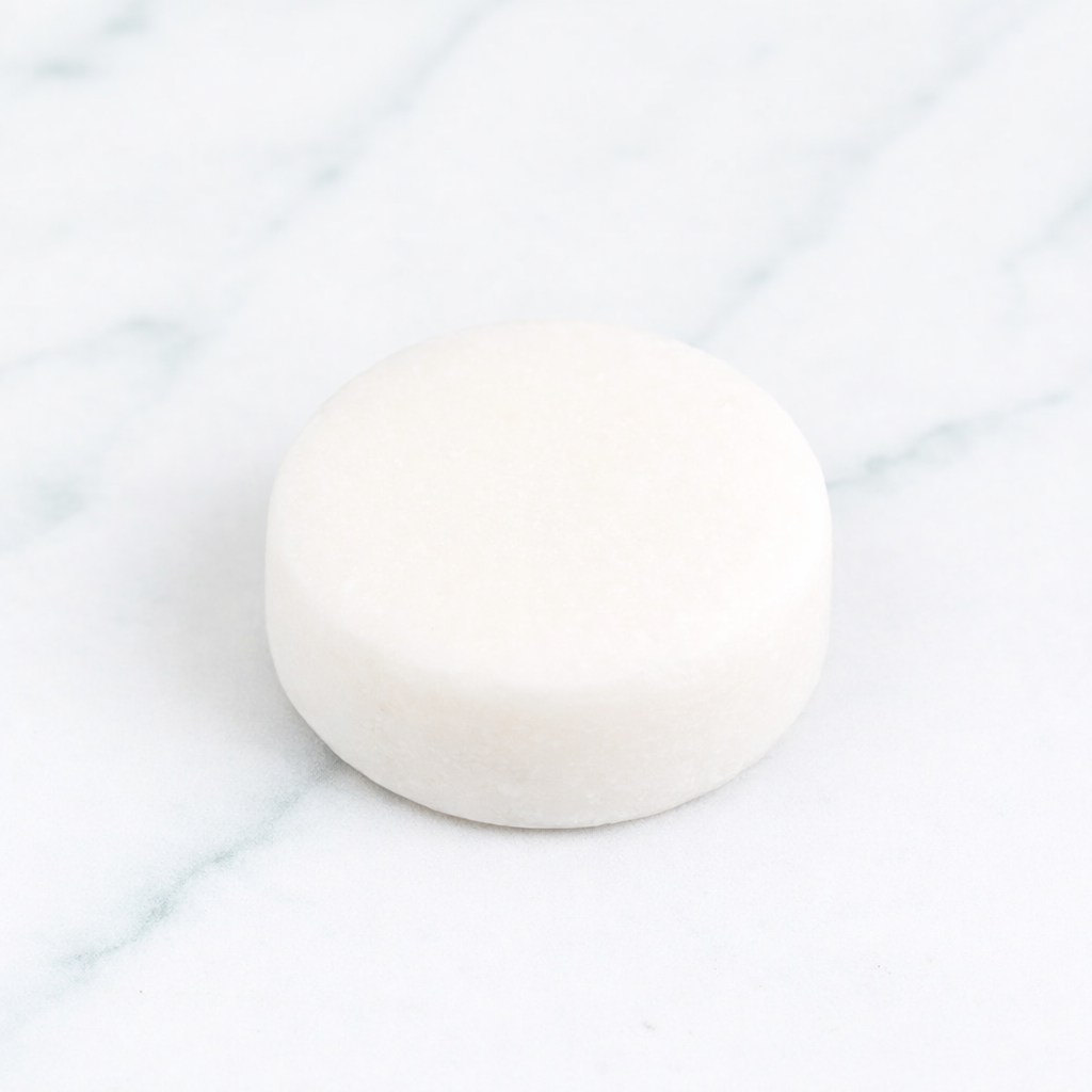A top-down professional shot of a round, white Citrus Mint Conditioner Bar with embossed lettering, centered on a white marble surface and surrounded by fresh orange slices, lemon halves, and vibrant mint leaves