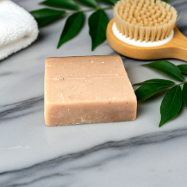tan, rectangular best bar soap for women displayed on a marble surface next to a wooden scrub brush and green leaves.