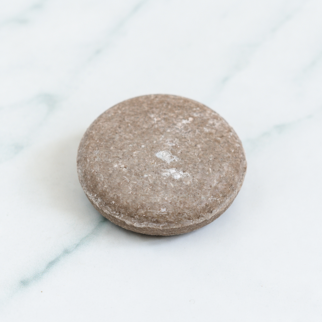 A professional close-up of a textured, earth-toned Rosemary Shampoo Bar. The bar is shown on a clean marble background, accompanied by fresh rosemary leaves and a wooden handle brush to emphasize its natural, stimulating ingredients.