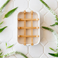 A white bar soap holder with a natural bamboo grid insert, displayed on a white hexagonal tile background with green botanical accents.