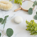 A round, white solid shampoo bar centered on a marble surface, near fresh eucalyptus leaves for an eco-friendly look.